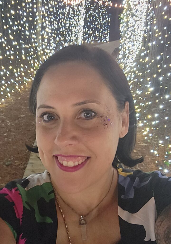 Selfie of Maegan Morrow, white woman smiling with shoulder-length brown hair and holiday lights in the background